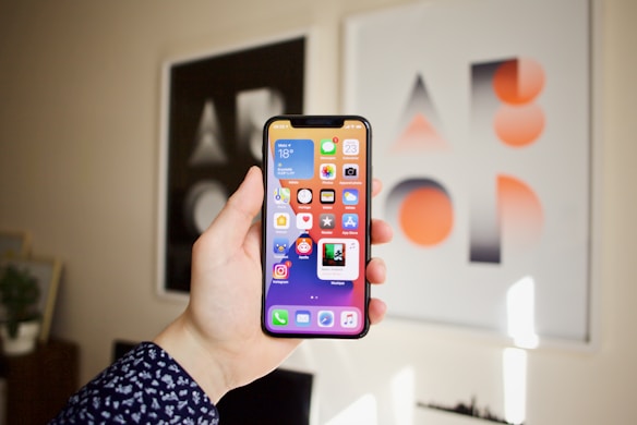 A hand is holding a smartphone displaying various app icons on the home screen. The background features two framed abstract art pieces with geometric designs in black, white, and orange.