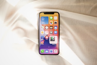 A smartphone displaying its home screen is placed on a softly wrinkled, light-colored fabric. The screen shows several app icons against a gradient background.
