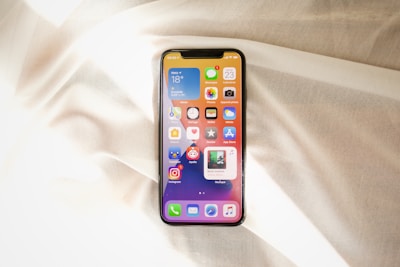 A smartphone displaying its home screen is placed on a softly wrinkled, light-colored fabric. The screen shows several app icons against a gradient background.