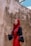 A person wearing a red hijab and a matching long skirt stands against a weathered and textured wall. The individual is dressed in a black top with loose sleeves, and the background features a sky with some clouds peeking over the wall.
