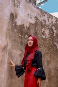 A person wearing a red hijab and a matching long skirt stands against a weathered and textured wall. The individual is dressed in a black top with loose sleeves, and the background features a sky with some clouds peeking over the wall.