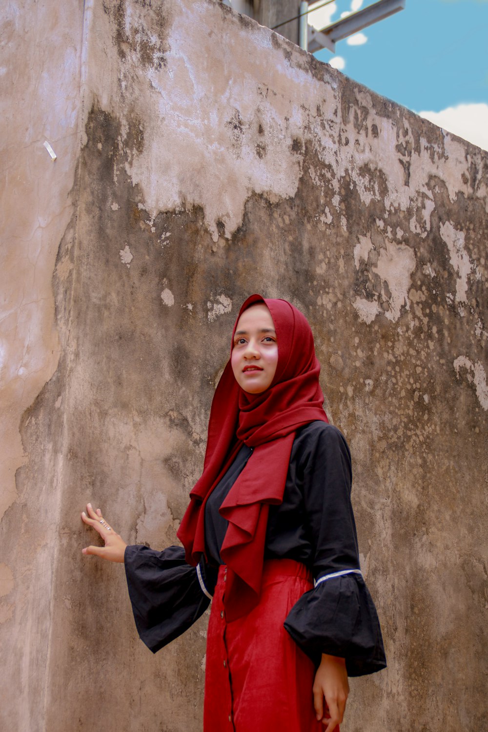 woman in red hijab and black long sleeve dress sitting on brown concrete wall