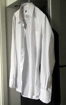 A neatly ironed white dress shirt hangs on a hanger against a neutral background. The shirt is buttoned up with long sleeves, a collar, and a front pocket.