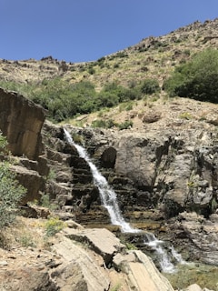 A small waterfall cascades down rugged rock formations surrounded by sparse vegetation. The hillside is dotted with green shrubs and bushes under a clear blue sky.