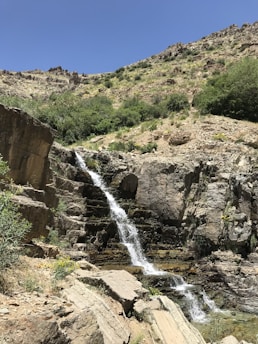 A small waterfall cascades down rugged rock formations surrounded by sparse vegetation. The hillside is dotted with green shrubs and bushes under a clear blue sky.