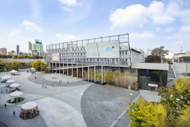 A modern outdoor plaza with a large, contemporary building featuring glass and metal elements. The plaza is paved with light gray tiles and has several small tents and people scattered around. The building is adorned with vegetation along its base and a digital display on its facade. Surrounding skyscrapers and trees are visible in the background.