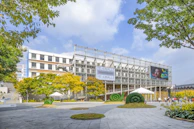 A modern building surrounded by lush green trees and landscaped gardens, featuring a prominent digital screen displaying a woman's face. The architecture includes large glass panels and a grid-like facade. There are several tents and well-maintained green topiaries in the foreground. The sky is partly cloudy with a clear blue background.