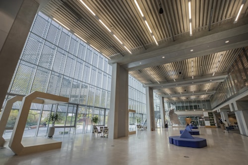 A spacious and modern interior with large windows letting in ample natural light. The architecture is characterized by a combination of glass and concrete, with an open atrium and multiple levels visible. Geometric designs, including a large letter 'C' structure and triangular seating, are featured throughout. Few plants and minimalist furniture add to the aesthetic.
