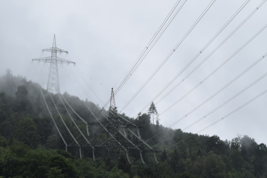 High-voltage power lines stretch across a misty, forested hillside. The tall transmission towers rise above the dense greenery, shrouded in fog that adds a mysterious atmosphere to the scene. The cables create a linear pattern against the overcast sky, emphasizing the engineering amidst natural surroundings.
