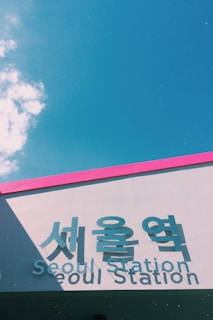 A modern building facade featuring large signage with both Korean and English text that reads 'Seoul Station'. The structure has a pink border against a clear blue sky, with some scattered clouds.