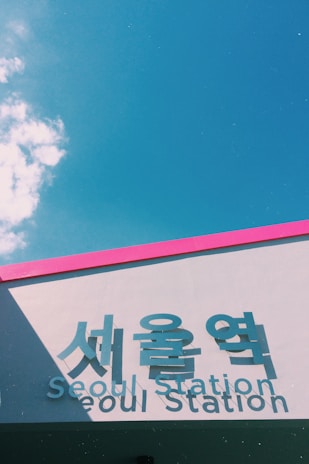 A modern building facade featuring large signage with both Korean and English text that reads 'Seoul Station'. The structure has a pink border against a clear blue sky, with some scattered clouds.