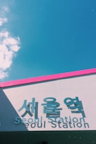 A modern building facade featuring large signage with both Korean and English text that reads 'Seoul Station'. The structure has a pink border against a clear blue sky, with some scattered clouds.