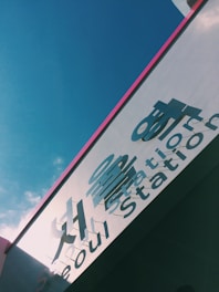 A sign for Seoul Station is captured from an upward angle, against a backdrop of a clear blue sky. The sign features large letters in both English and Korean, casting shadows against the surface. A hint of pink framing adds a touch of color.