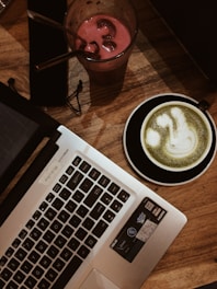 A laptop is positioned on a wooden table, accompanied by two drinks: a matcha latte on the right in a black cup and saucer, and a pink smoothie with ice and a straw in a clear glass on the left. The scene conveys a cozy cafe vibe with a focus on work or relaxation.