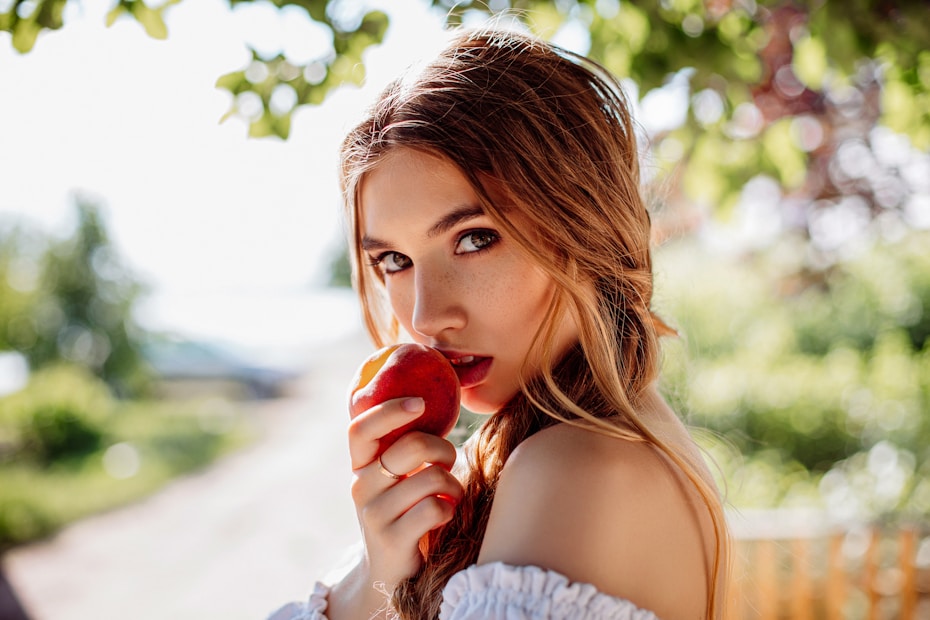 A woman with long hair is holding a red apple close to her mouth. She is surrounded by lush greenery and soft sunlight filters through the leaves, creating a light and airy atmosphere.