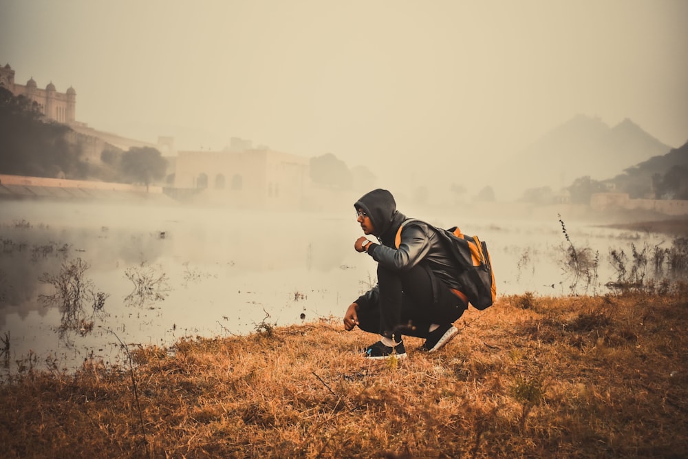 man in black jacket and black pants sitting on brown grass field near lake during daytime