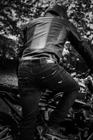 A person wearing a leather jacket and helmet is sitting on a motorcycle. The image is in black and white, giving it a classic and timeless feel. The motorcycle has detailed mechanical parts visible, and the background consists of blurred trees, suggesting a natural setting.