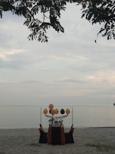 A solitary birthday setup on a sandy beach with a table adorned in black and orange fabric. Balloons are arranged above the table with a 'Happy Birthday' sign, and leaves from a tree partially frame the top of the scene, with a calm sea in the background.
