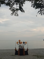 A solitary birthday setup on a sandy beach with a table adorned in black and orange fabric. Balloons are arranged above the table with a 'Happy Birthday' sign, and leaves from a tree partially frame the top of the scene, with a calm sea in the background.