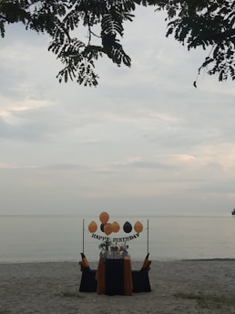 A solitary birthday setup on a sandy beach with a table adorned in black and orange fabric. Balloons are arranged above the table with a 'Happy Birthday' sign, and leaves from a tree partially frame the top of the scene, with a calm sea in the background.