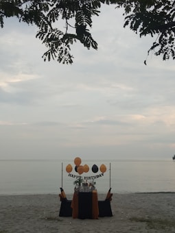 A solitary birthday setup on a sandy beach with a table adorned in black and orange fabric. Balloons are arranged above the table with a 'Happy Birthday' sign, and leaves from a tree partially frame the top of the scene, with a calm sea in the background.