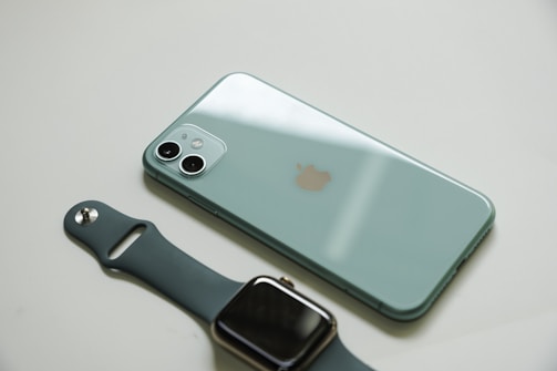 A mint green smartphone with a dual-camera system is positioned on a light surface, adjacent to a dark-colored smartwatch with a metallic buckle.