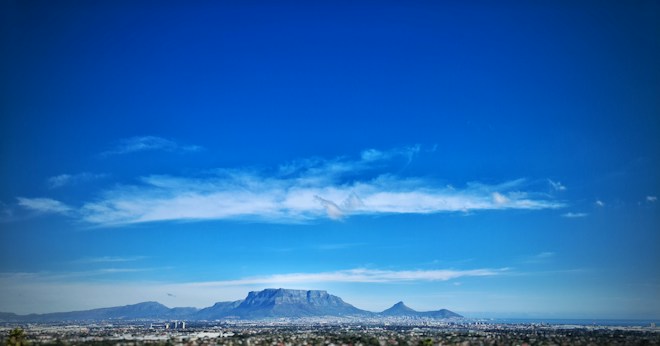 A vast clear blue sky with a few wispy clouds scattered across it. Below, a mountain range is visible with the prominent flat-topped Table Mountain in the center and city buildings spread out at the base. The landscape is predominantly green and brown with some urban development.