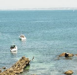 Three small boats float on a vast blue ocean, with one boat closer in the foreground and two further in the distance. A person is seen mid-jump off a large rock into the water, suggesting leisure and adventure. The distant shoreline is visible on the horizon under a clear sky.