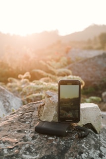 A smartphone is placed on a rock in a natural outdoor setting, with the screen displaying the same landscape that is in the background. The scene is bathed in warm, golden sunlight creating a serene and peaceful atmosphere.  Surrounding the setup are large rocks and some greenery.