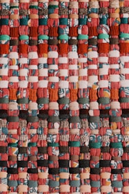 A colorful, tightly woven textile pattern composed of alternating bands of various fabrics. The materials are intricately interlaced, featuring warm hues like reds, oranges, and pinks with accents of teal and black. The texture appears rich and tactile, indicative of handcrafted artistry.