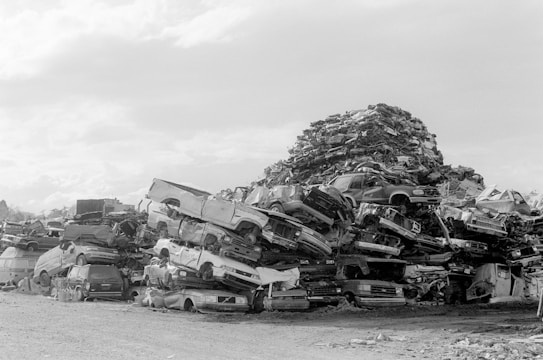 A large pile of crushed and stacked vehicles forms a chaotic mountain in what appears to be a junkyard or scrapyard. The cars are mostly stripped and stacked haphazardly on top of one another against a clear sky.