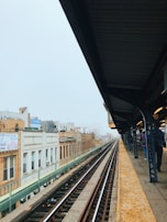An elevated train platform with rail tracks running through an urban environment. Buildings with signs in different languages are visible on the left side. The platform has yellow tactile paving, and a few people are waiting with their backs turned.