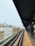 An elevated train platform with rail tracks running through an urban environment. Buildings with signs in different languages are visible on the left side. The platform has yellow tactile paving, and a few people are waiting with their backs turned.
