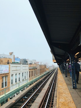 An elevated train platform with rail tracks running through an urban environment. Buildings with signs in different languages are visible on the left side. The platform has yellow tactile paving, and a few people are waiting with their backs turned.