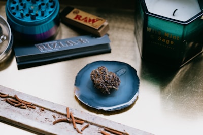 A collection of items placed on a smooth surface includes a herb grinder, rolling papers with 'RAW' branding, a candle labeled 'Wild Mint & Sage', and a decorative crystal dish holding a small piece of dried herb or plant material. An incense stick with ashes is also present.