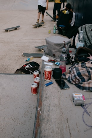 Several skateboards rest on a concrete surface alongside a group of young people, some standing and others crouching. Various items are scattered on a ramp, including soda cans, a smartphone, water bottles, a lighter, and a pack of cigarettes. A patterned shirt and a gray backpack add color and texture to the scene. The setting appears to be an informal outdoor gathering, possibly at a skatepark.