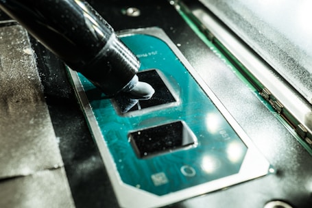 A close-up view of a CPU socket on a motherboard with a thermal paste being applied using an application tube. The components are part of computer hardware, with the metallic socket reflecting light.