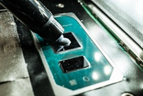 A close-up view of a CPU socket on a motherboard with a thermal paste being applied using an application tube. The components are part of computer hardware, with the metallic socket reflecting light.