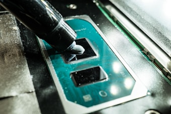 A close-up view of a CPU socket on a motherboard with a thermal paste being applied using an application tube. The components are part of computer hardware, with the metallic socket reflecting light.
