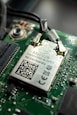 Close-up view of a computer circuit board featuring a Wi-Fi module with labels and a QR code. The module is connected to the board with visible soldered connections and surrounding electronic components.