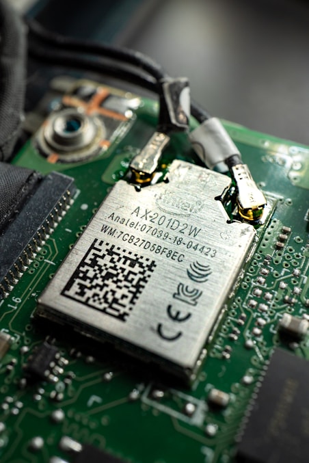 Close-up view of a computer circuit board featuring a Wi-Fi module with labels and a QR code. The module is connected to the board with visible soldered connections and surrounding electronic components.