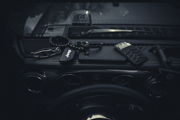 Inside a car, the dashboard features various items such as a Jeep key fob, a multi-tool, a wristband, a flashlight, and a wallet. The lighting is dim, creating a moody atmosphere.