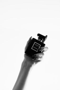 A monochrome image features a hand with neatly manicured nails holding a perfume bottle labelled 'Coco Chanel'. The composition is minimalist, focusing on the elegant design of the bottle contrasted against a plain white background.