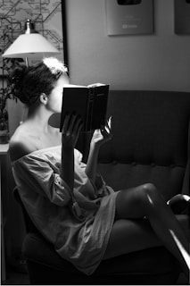 A person sits comfortably on a chair, engrossed in reading a book. The light casts an artistic shadow across the person's face and body, creating a tranquil and intimate atmosphere. The room includes a lamp, a map on the wall, and plaques that suggest a creative or literary setting.