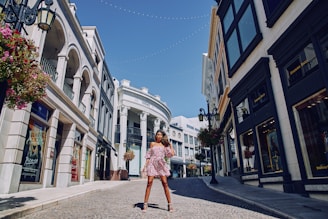 A woman is standing confidently in the middle of a cobblestone street lined with upscale shops and buildings featuring classical architecture. She is wearing a floral off-the-shoulder dress and high heels. There are ornate hanging lanterns and potted flowers adorning the street.