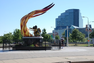 A large, dynamic sculpture of a firefighter with vibrant, flowing flames is positioned prominently in a public space. The sculpture is surrounded by a black metal fence, landscaping, and is set against a backdrop of modern multi-story glass buildings. Nearby, a person dressed in traditional Scottish attire, including a kilt, is playing the bagpipes.