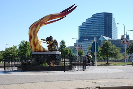 A large, dynamic sculpture of a firefighter with vibrant, flowing flames is positioned prominently in a public space. The sculpture is surrounded by a black metal fence, landscaping, and is set against a backdrop of modern multi-story glass buildings. Nearby, a person dressed in traditional Scottish attire, including a kilt, is playing the bagpipes.