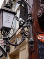 A decorated hanging sign with ornate metallic designs and a lantern projecting from a building, which advertises 'The Real Dutch Pub Experience.' A string of decorative lights is wrapped around parts of the sign and building. To the right, there is a blackboard with partial text and red signage. The building facade features ornate architectural details and elements in beige and brown colors.