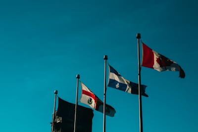Four flags are raised on tall poles against a clear sky. The flags include the French Canadian flag and another with a red maple leaf.