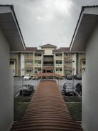 A residential area features a row of yellow apartment buildings with brown tiled roofs. There is a covered walkway leading towards the buildings, surrounded by parked cars and metal lamp posts. The sky is overcast, suggesting cloudy weather.